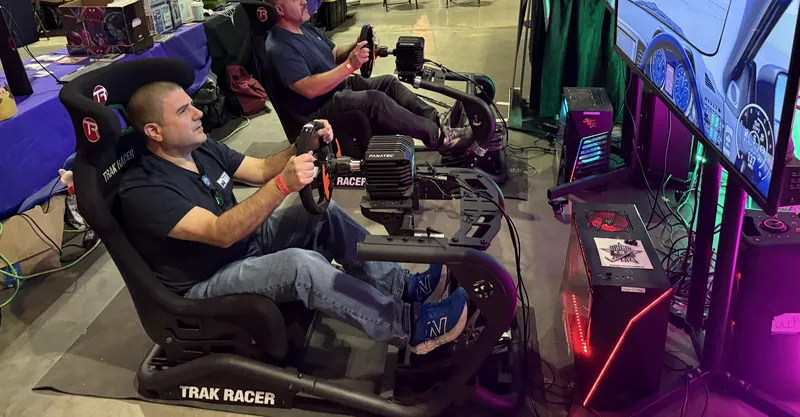 A driver in a full racing simulator rig, hands on the wheel, racing on a big screen at a Next Level Gaming event.